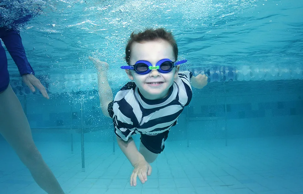 kleiner Junge ist gerade am Tauchen. Er hat eine blaue Taucherbrille auf und ein gestreiftes Schwimm-Oberteil an.