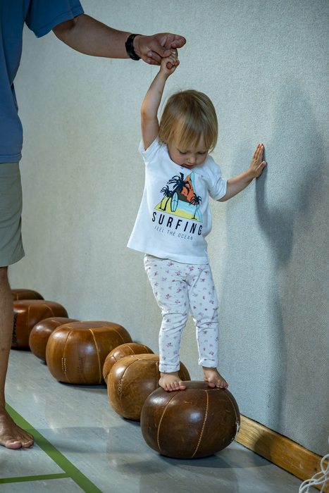 kleines Mädchen balanciert auf Medizinbällen und stützt sich mit einer Hand an der Turnsaalwand an und mit der anderen Hand hält sie sich an einer anderen Hand fest.